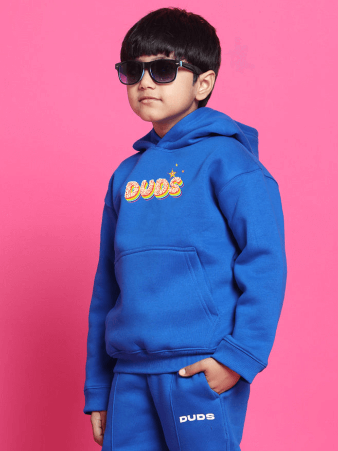Child wearing blue oversized 'Born to Shine' hoodie and sunglasses, posing confidently against a pink backdrop.