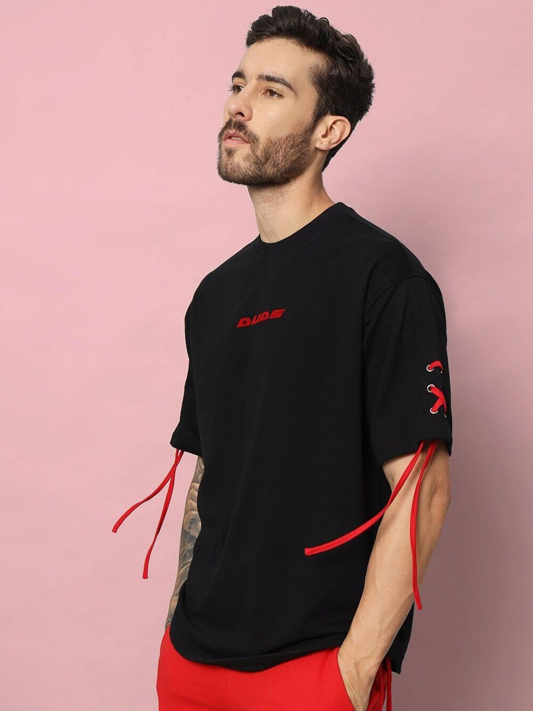 Man wearing BOWLINE OVER-SIZED T-SHIRT BLACK with red accents, standing against a pink background, showcasing casual style.