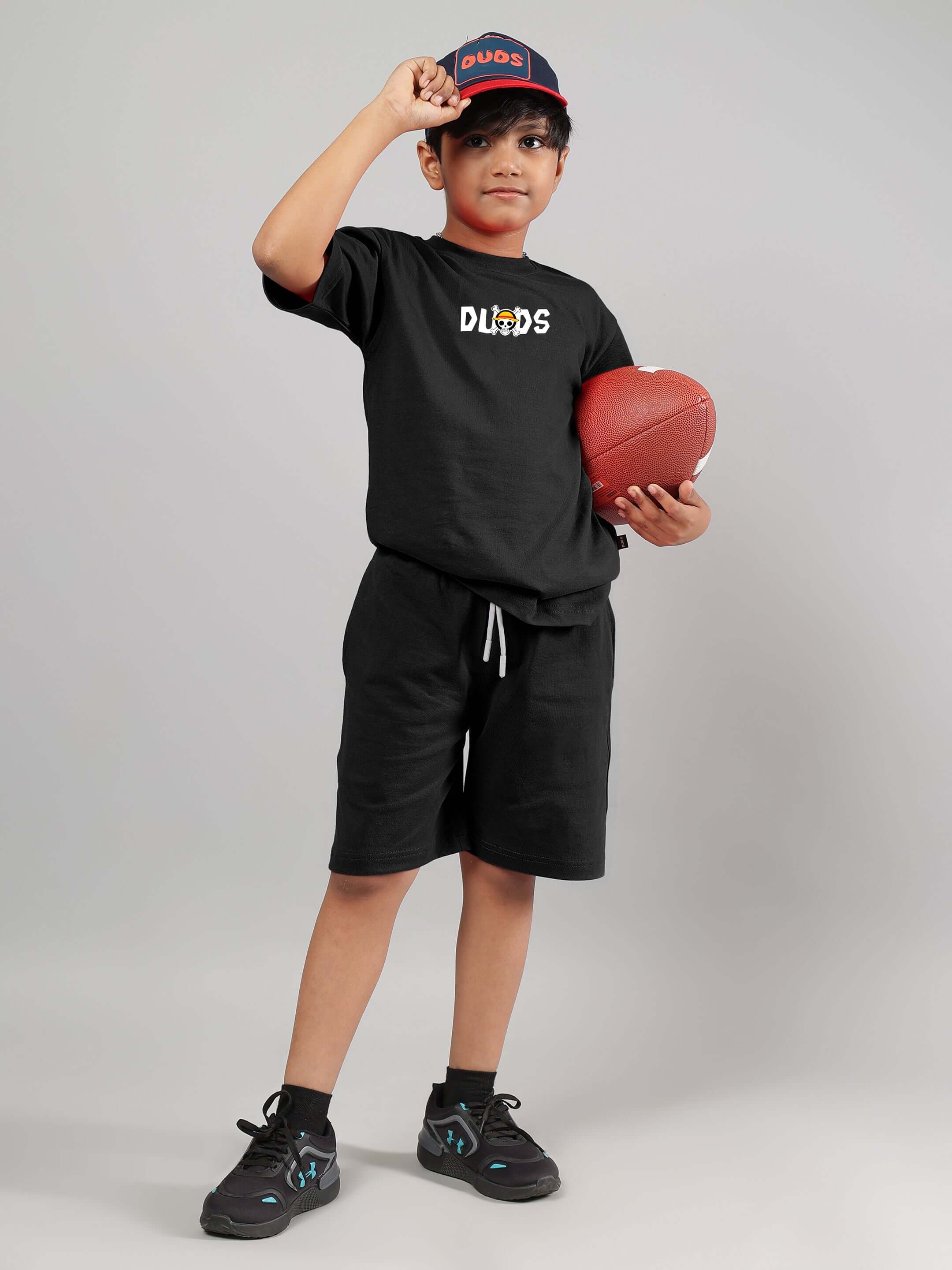 Boy wearing black ONE PIECE T-shirt holding a football, dressed in shorts and cap by DUDS, showcasing stylish casual wear for kids.