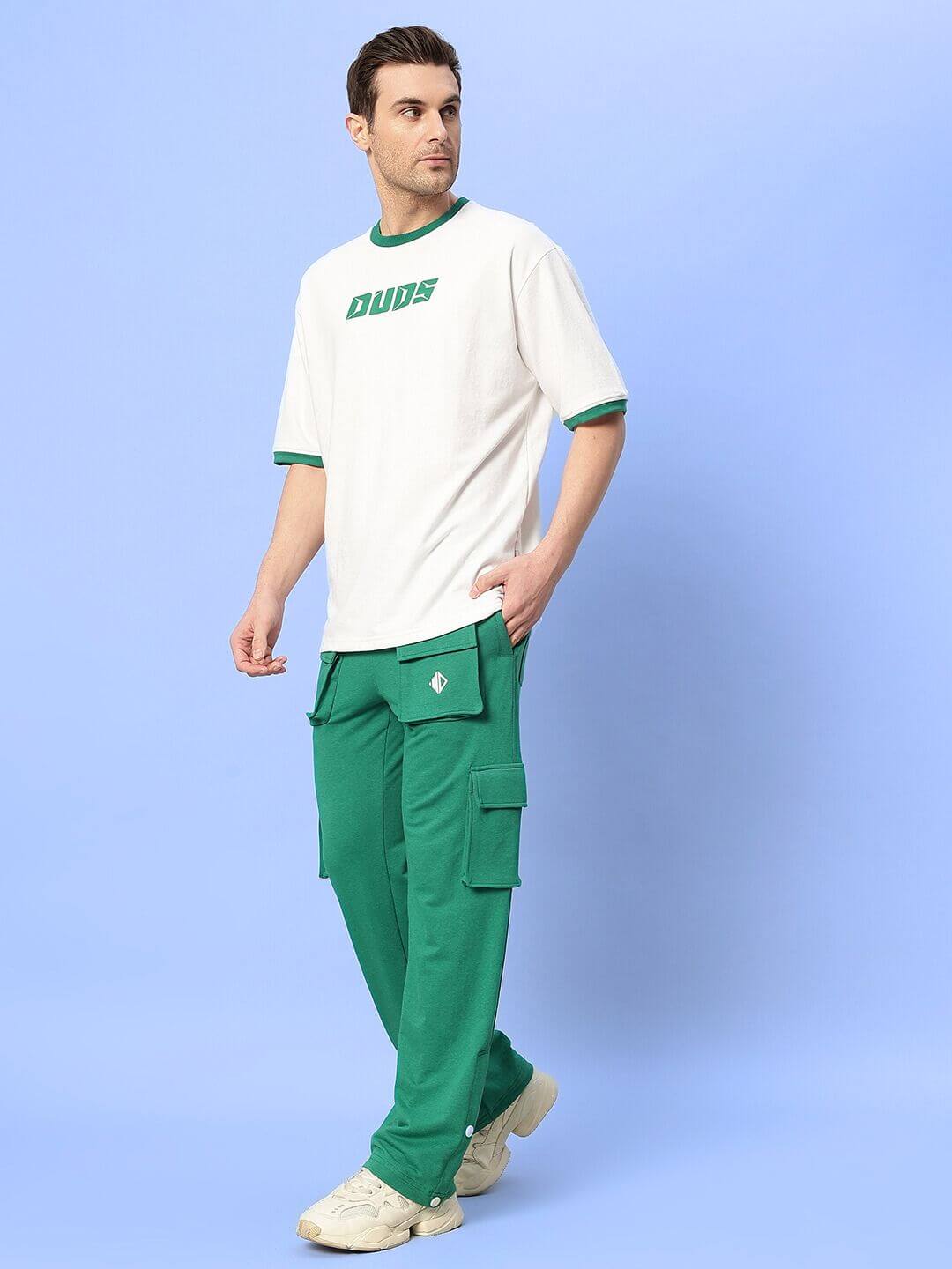 Man wearing teal green cargo pants with multiple pockets, paired with a white DUDS t-shirt and white sneakers, standing against a blue background.