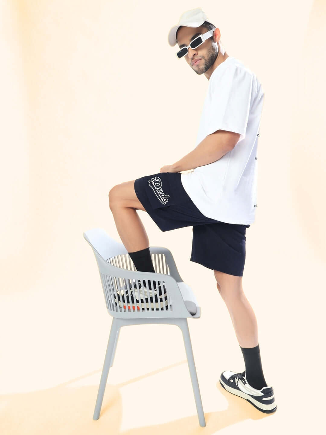 Man wearing navy blue Brawny cargo shorts and white t-shirt with sunglasses, standing on a chair for a casual style photoshoot.