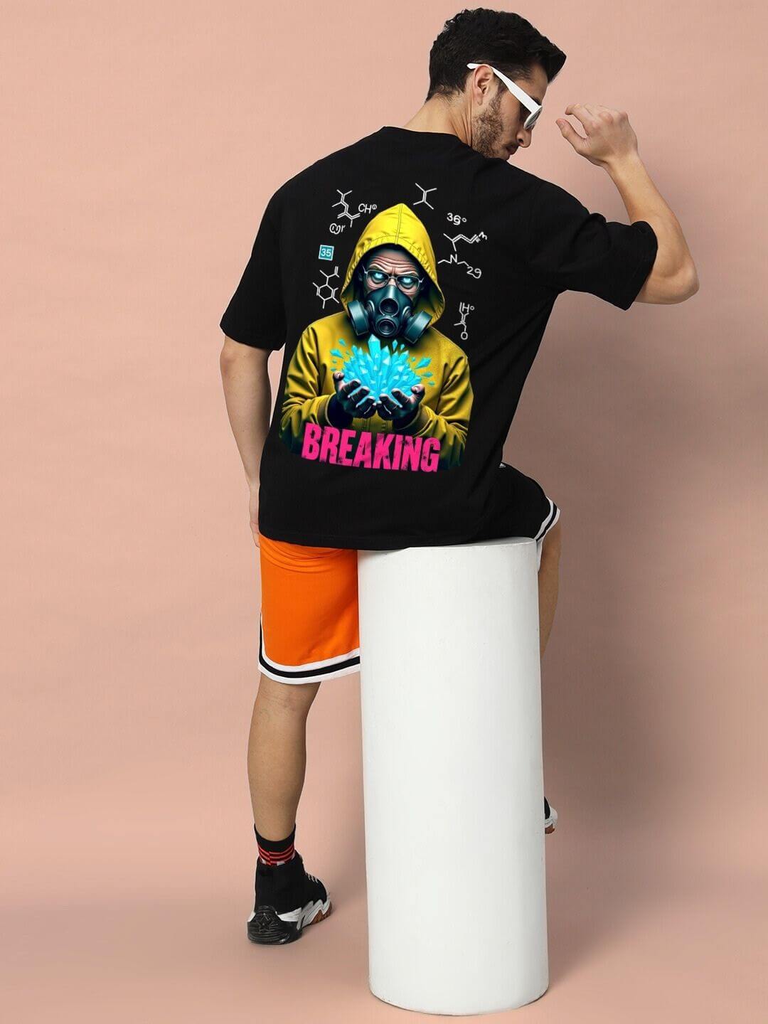 Model wearing BREAKING OVER-SIZED T-SHIRT BLACK with graphic back print, seated on a stool, showcasing urban streetwear style.