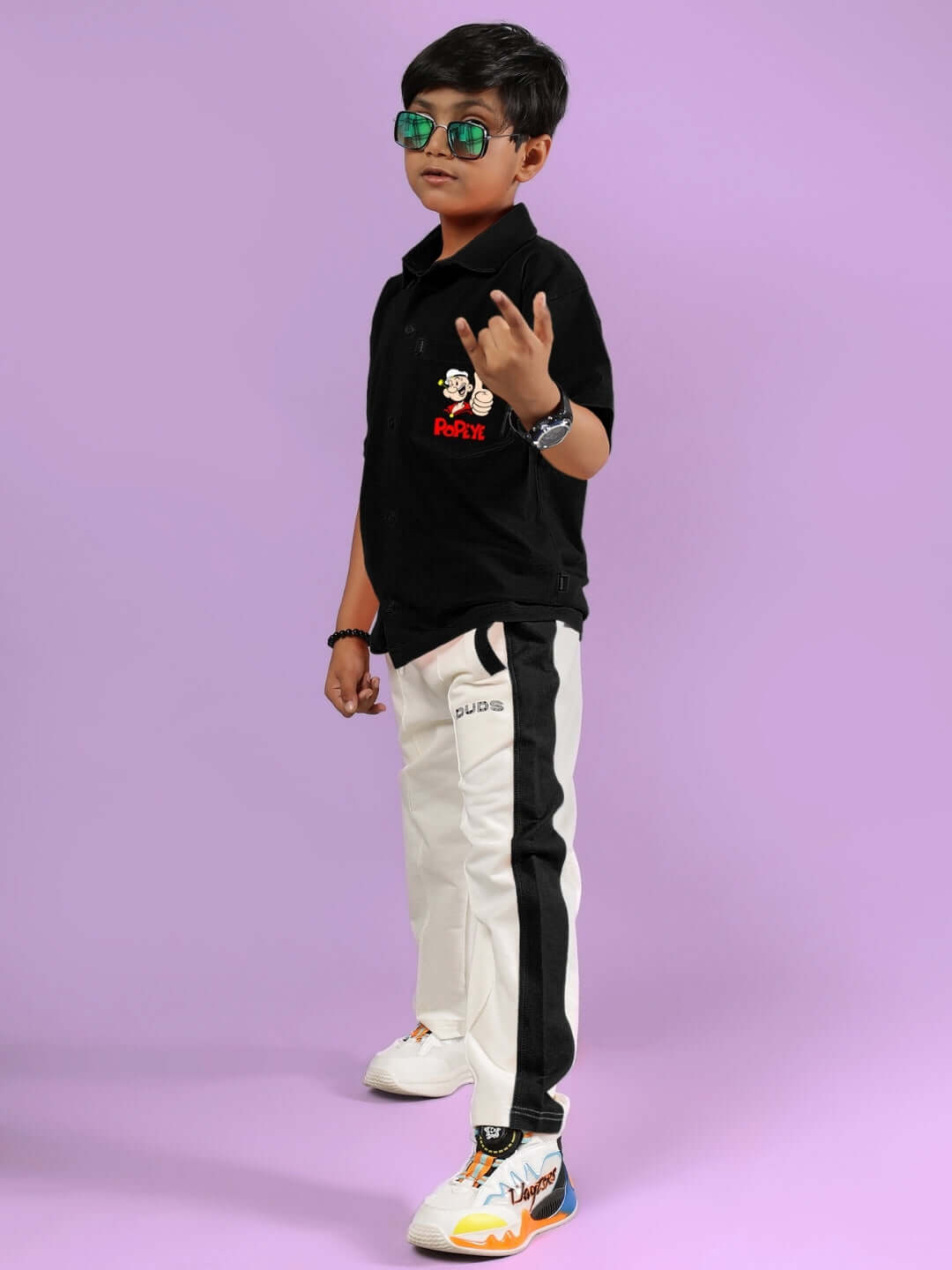 Young boy wearing DUDS off-white and black Breezer Jogger, with sunglasses and a black shirt, standing confidently against a purple background.