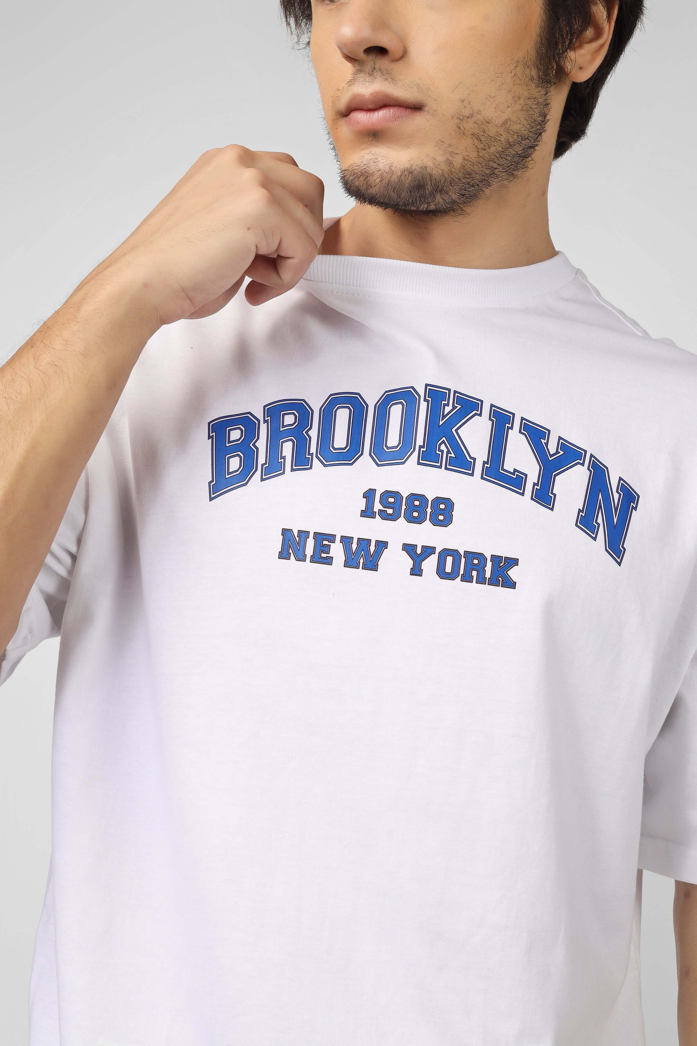 White t-shirt with 'BROOKLYN 1988 NEW YORK' blue print, worn by a model. Fashionable casual wear for contemporary style.