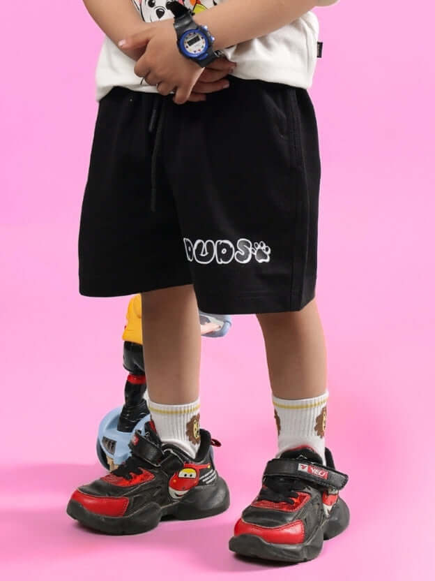 Child wearing BRUNO SHORTS FOR BOYS & GIRLS BLACK with cartoon shoes on pink background.