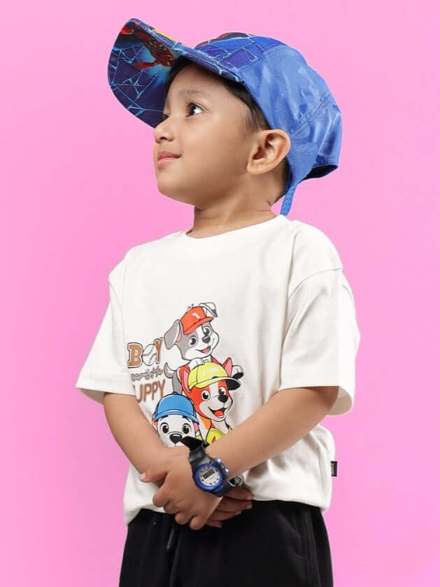 Child wearing BRUNO OVERSIZED T-SHIRT FOR BOYS GIRLS OFF WHITE with cartoon print, blue cap, and watch, standing against pink background.