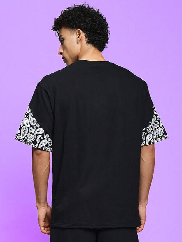 Model wearing CALVIN OVER-SIZED T-SHIRT BLACK with paisley sleeves, showcasing back design against purple background