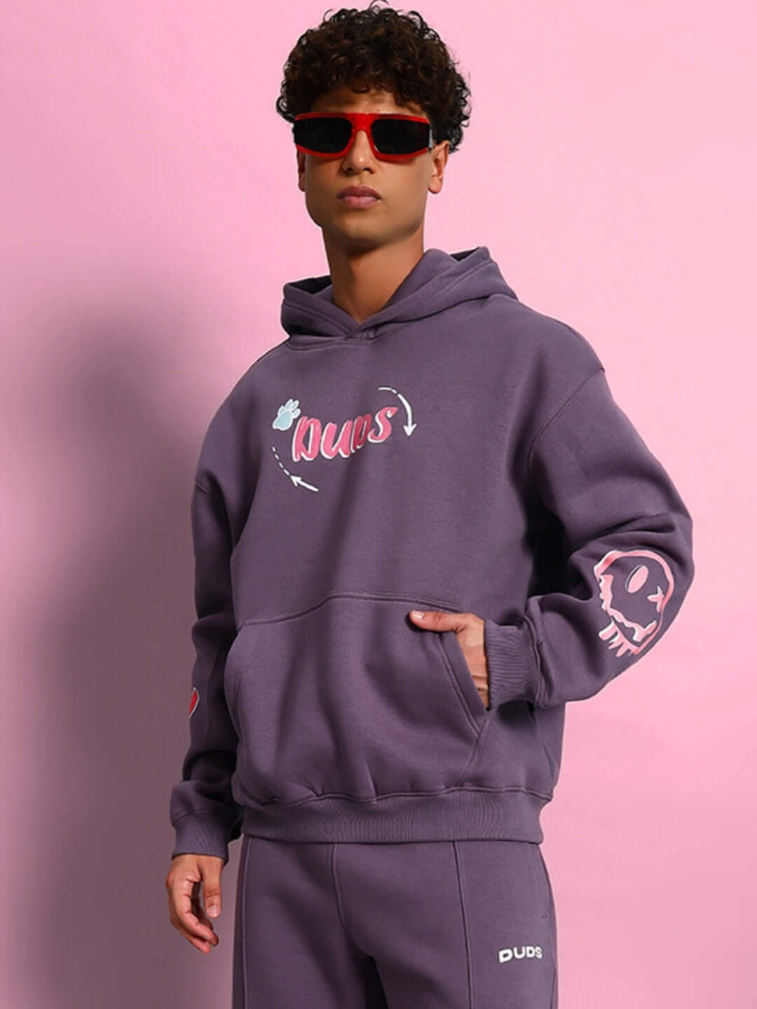 Model wearing CASPER COTTON FLEECE HOODIE VIOLET with red sunglasses, pink background.
