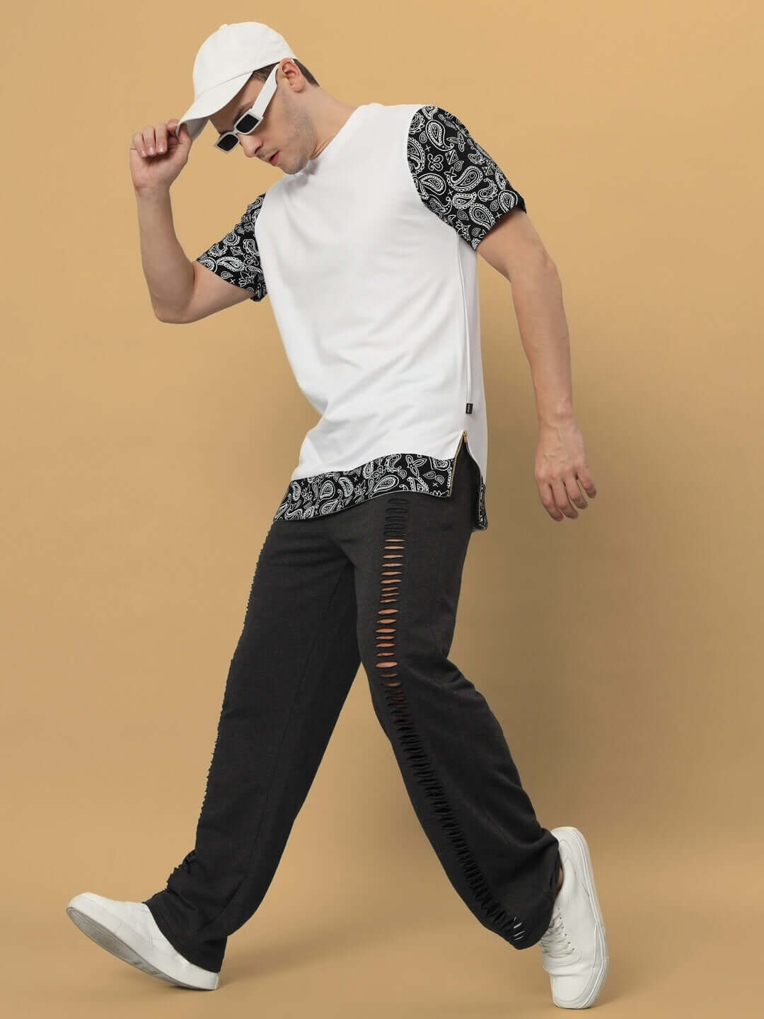 Man wearing stylish white cap, black patterned t-shirt, and white sneakers with shredded black pants against a beige background.