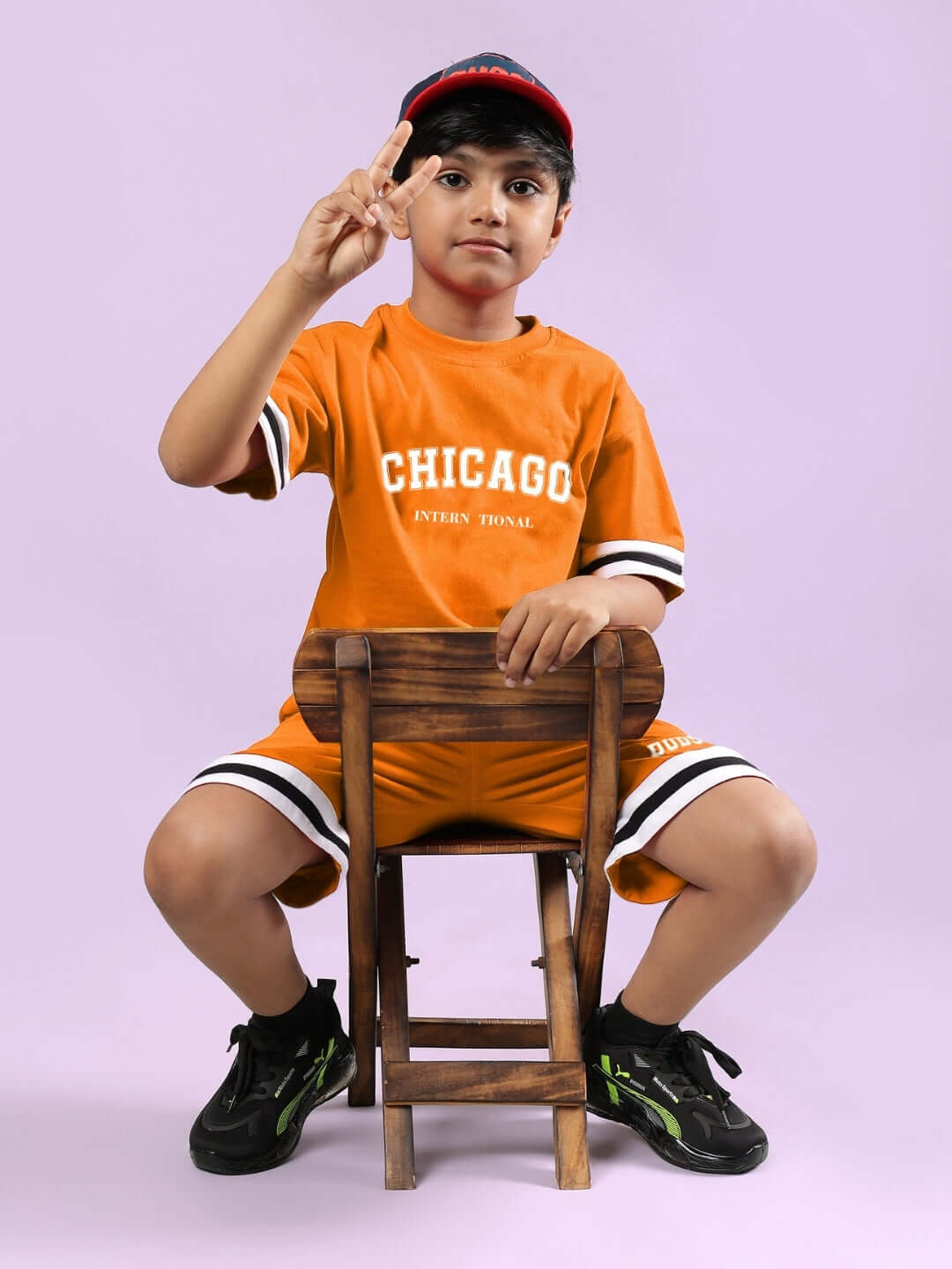 Child wearing CHICAGO PRINTED CO-ORD FOR BOYS & GIRLS ORANGE, sitting on a stool, peace sign, purple background.