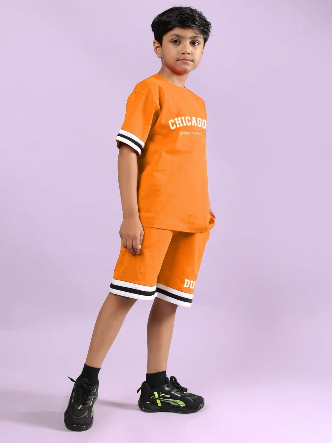 Child wearing CHICAGO PRINTED CO-ORD FOR BOYS & GIRLS ORANGE with sporty shoes, standing confidently against a purple background.