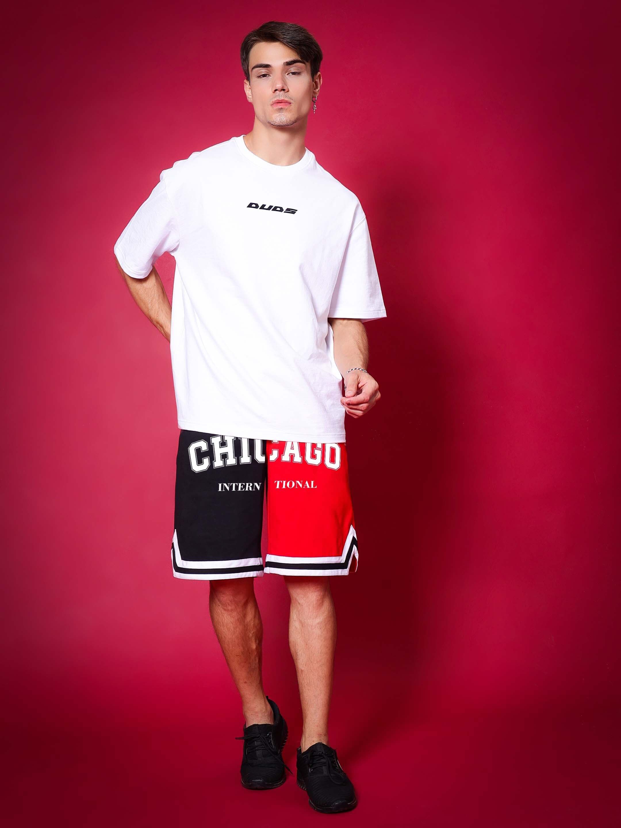 Model wearing Chicago Regular Fit Shorts Red Black with a white t-shirt, standing against a red background, showcasing casual style.