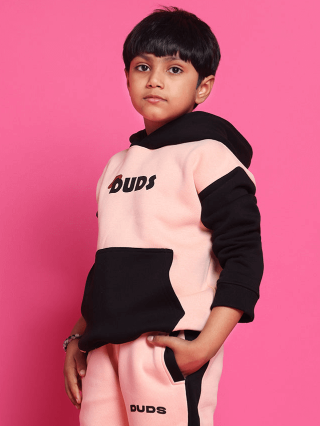 Child wearing a stylish pink and black hoodie with 'Duds' logo, standing confidently against a vibrant pink background.