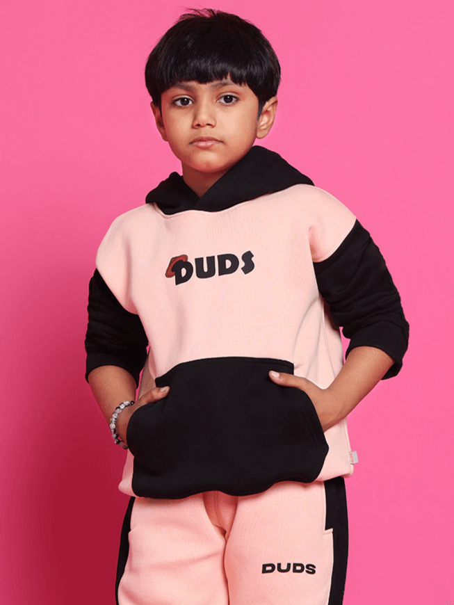 Child wearing a black-beige oversized hoodie with "Duds" logo, posing against a pink background.