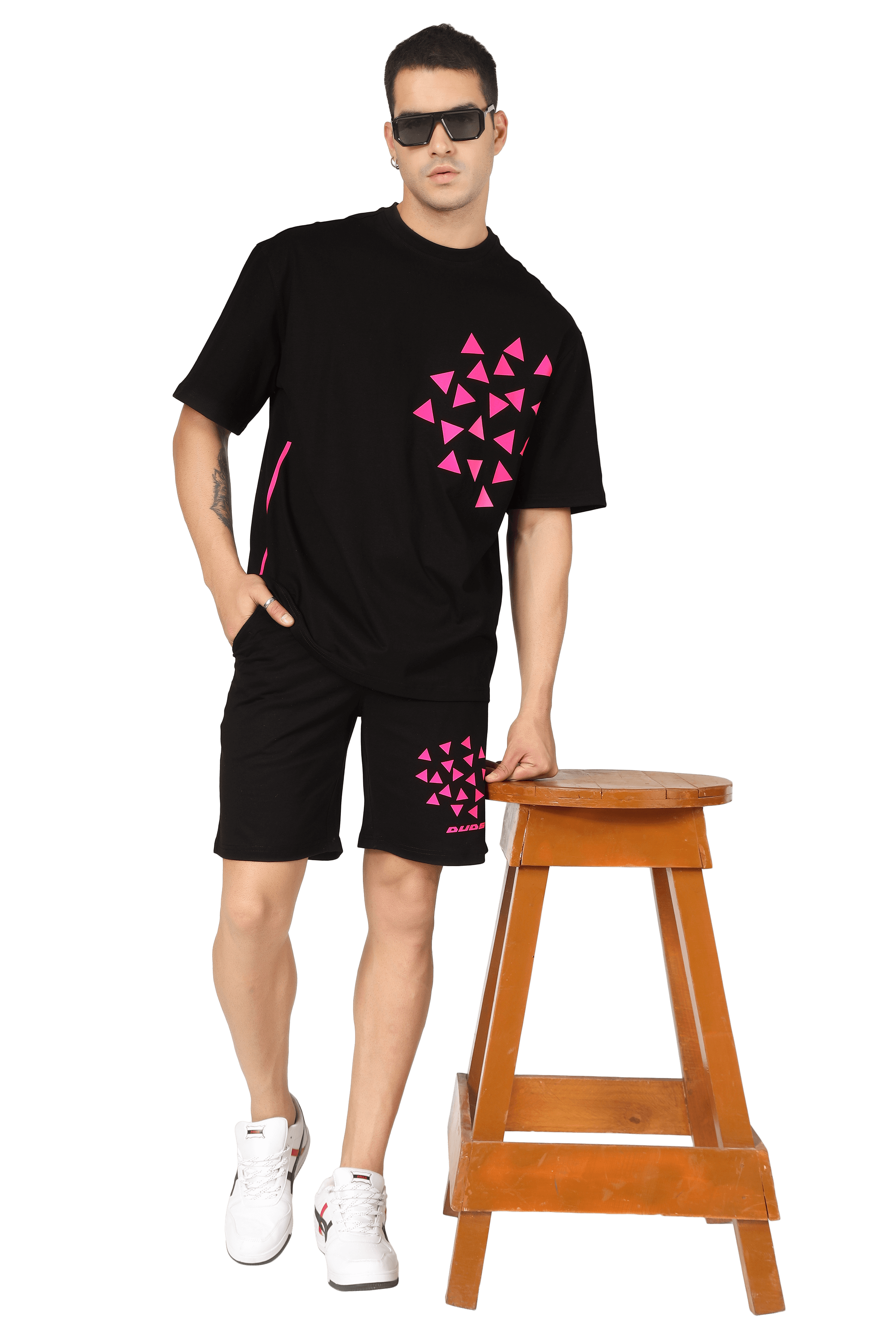 Model in black Star Dust CO-ORD set with pink geometric print, standing casually by a wooden stool, wearing sunglasses and white sneakers.