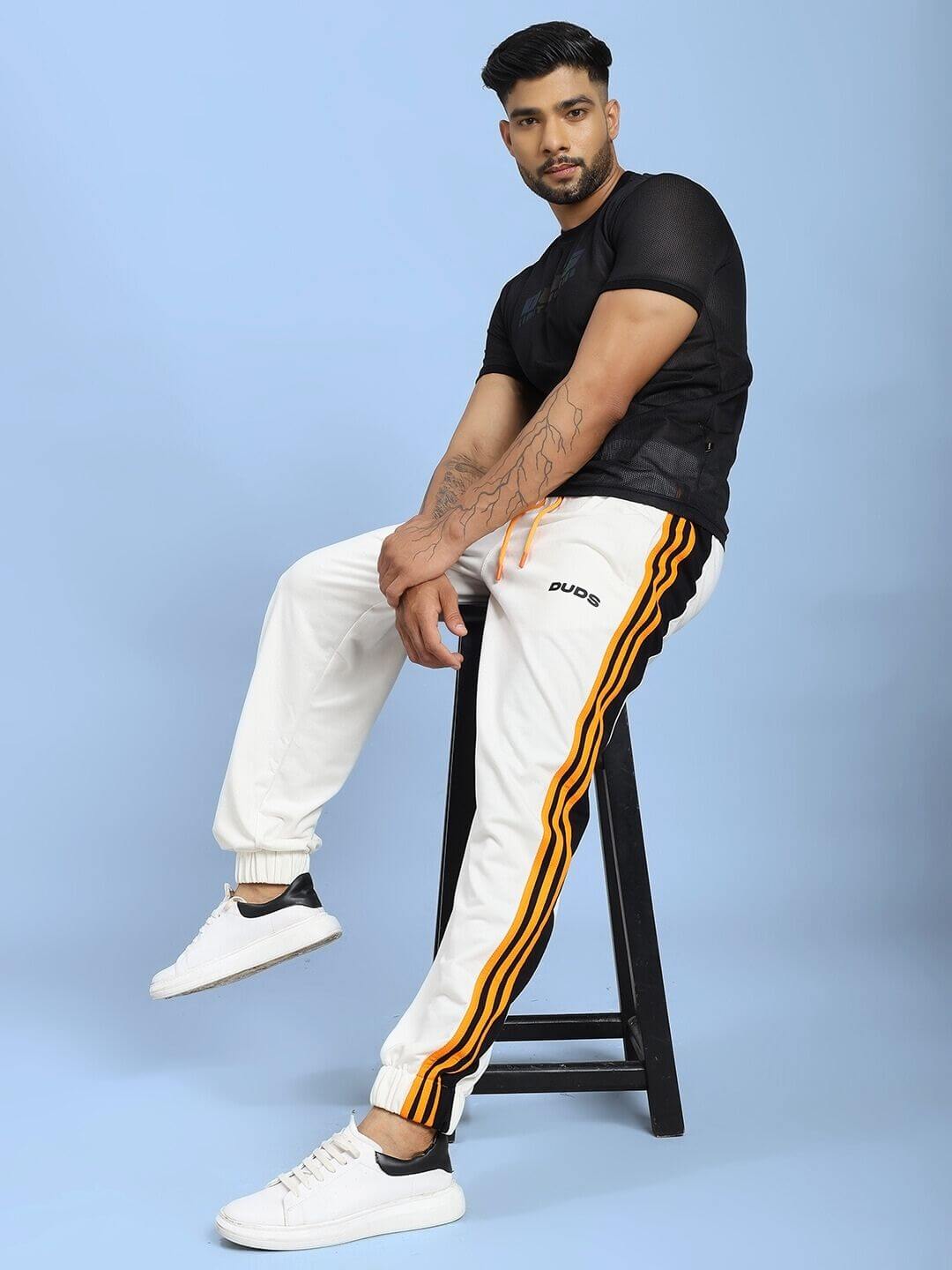 Man sitting on stool wearing COMFORT FLEX STRIPE JOGGER OFF-WHITE with orange stripes, black shirt, and white shoes against blue background.