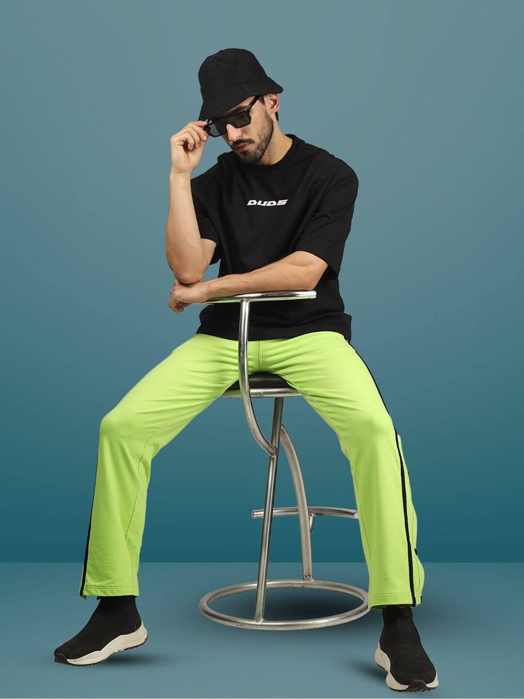 Man wearing neon green contrast panel joggers by DUDS, sitting on a stool against a blue background.