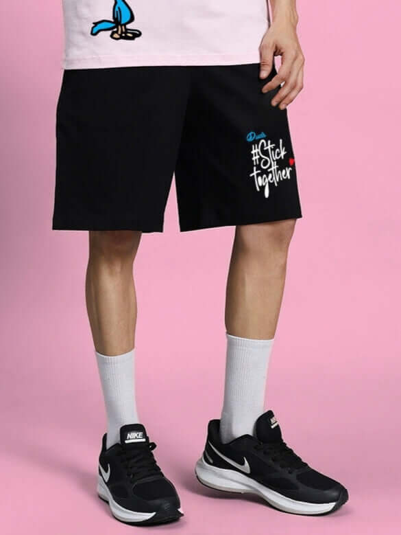 Model wearing COOL PALS REGULAR FIT SHORTS BLACK with hashtag print, showcasing casual style on a pink background.