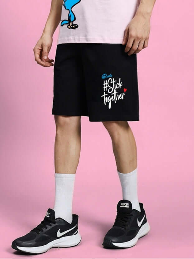 Model wearing COOL PALS REGULAR FIT SHORTS BLACK with graphic design, standing against a pink background, showcasing casual streetwear fashion.