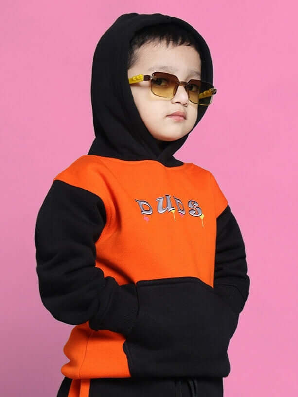 Child wearing orange-black oversized hoodie and sunglasses against pink background