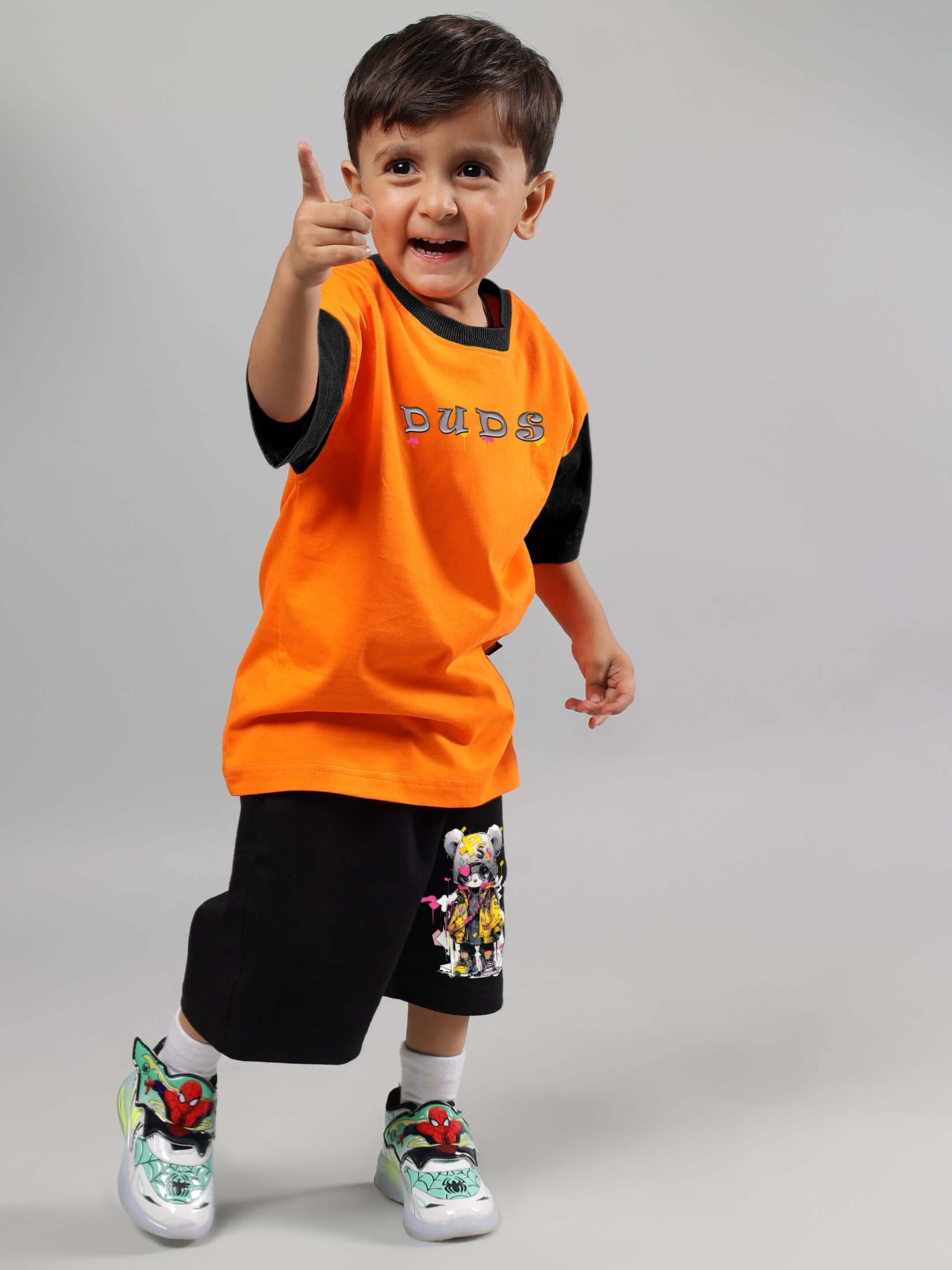 Child wearing black COOL TEDDY shorts and orange DUDS shirt, pointing excitedly, showcasing vibrant kids' fashion.