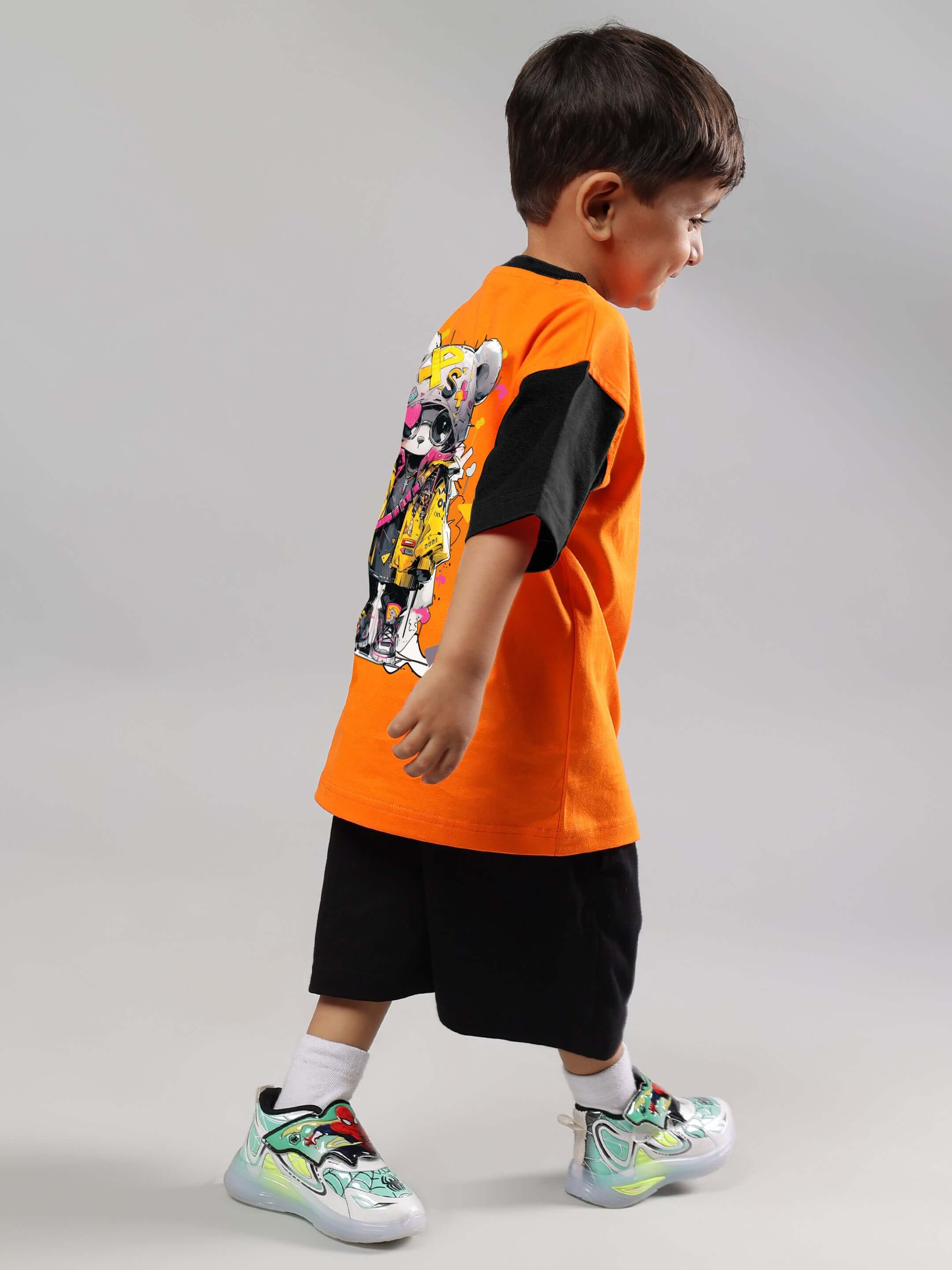 Boy wearing orange COOL TEDDY T-shirt for boys & girls from DUDS, side view with colorful design on back, paired with black shorts.