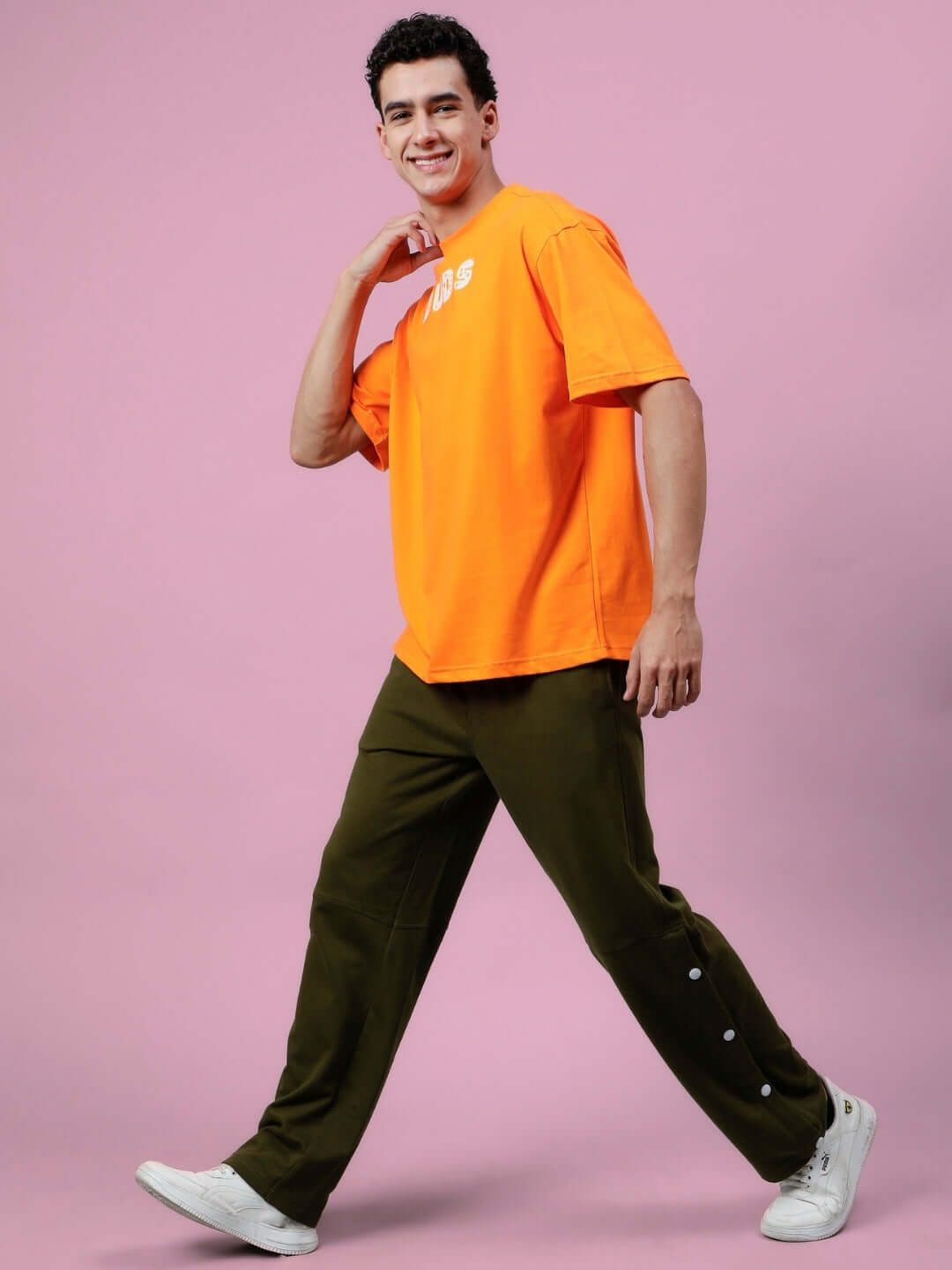 Man wearing orange COURAGE oversized t-shirt with olive pants, walking against a pink background.