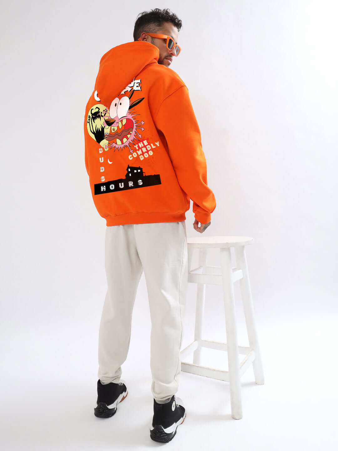 Person wearing an orange oversized hoodie with graphic design, standing next to a white stool.
