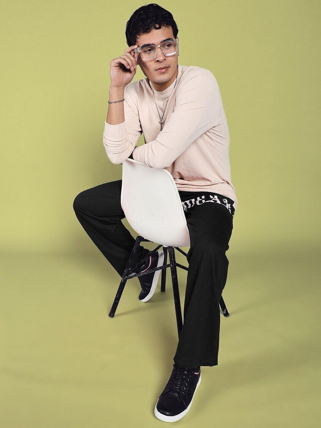 Man wearing black joggers and a beige sweater sitting on a chair against a green background.