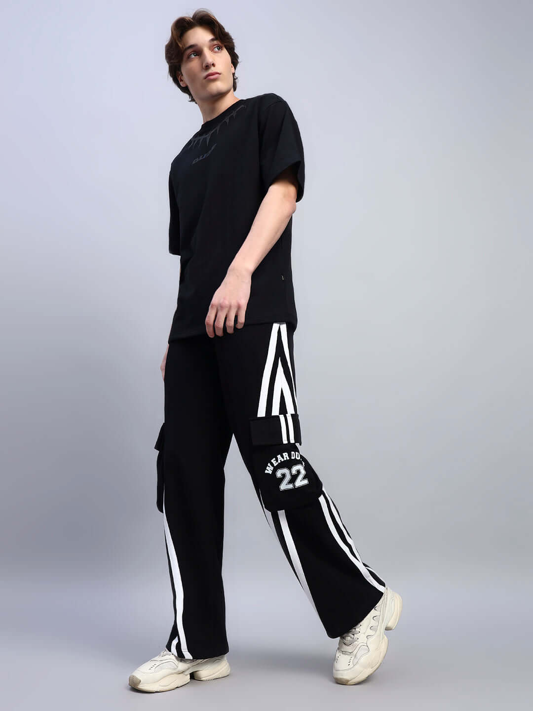 Model wearing black CRUCIS multi-pocket relaxed-fit joggers with white stripe design, paired with a black shirt and white sneakers.