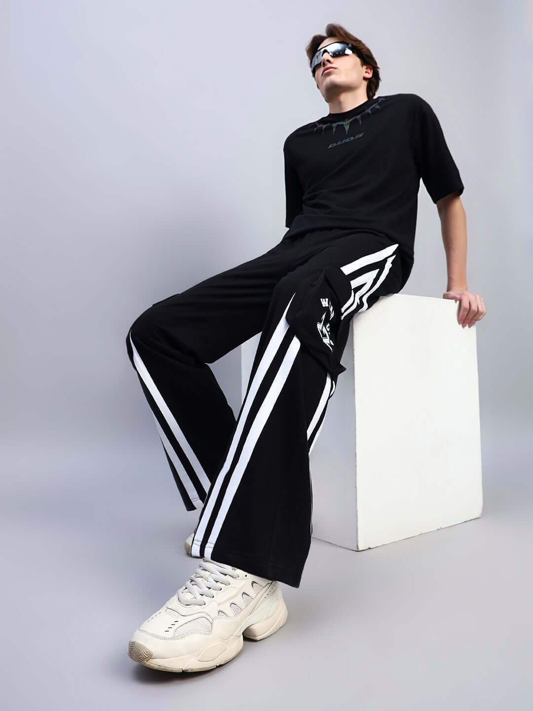 Man wearing black multi pocket relaxed-fit joggers with white sneakers sitting on white block, showcasing streetwear style.