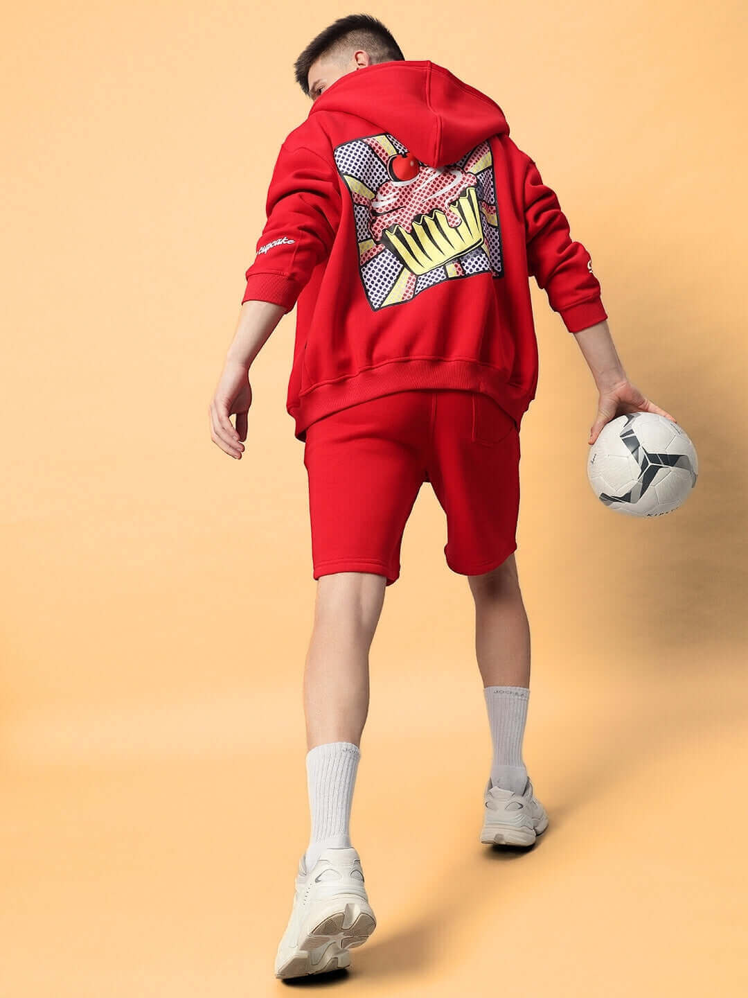 Young man wearing a vibrant red CUPCAKE COTTON FLEECE HOODIE RED holding a soccer ball. Trendy streetwear style.