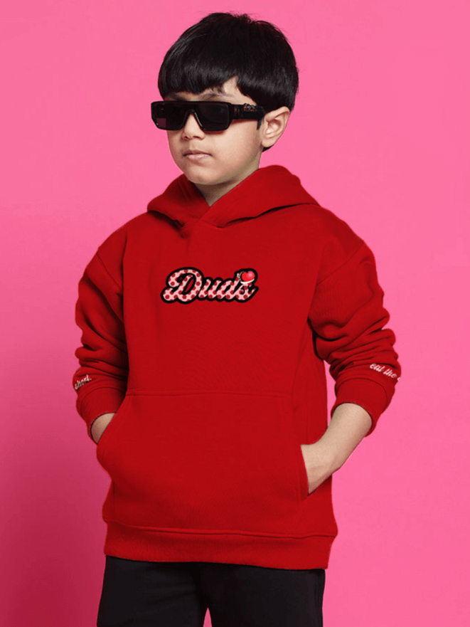 Child wearing a red oversized hoodie with "Dude" design, hood up, sunglasses, against a pink background.