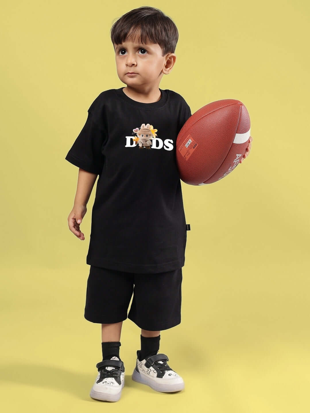 Young child in black 'CUTE MONSTERS CO-ORD' outfit holding a football, showcasing stylish kids' clothing by DUDS.