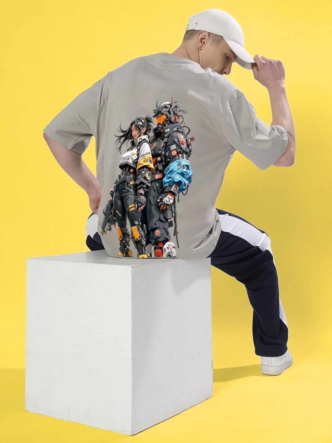 Man wearing CYBERPUNK OVER-SIZED T-SHIRT GREY with futuristic design, sitting on a white cube against a yellow background.