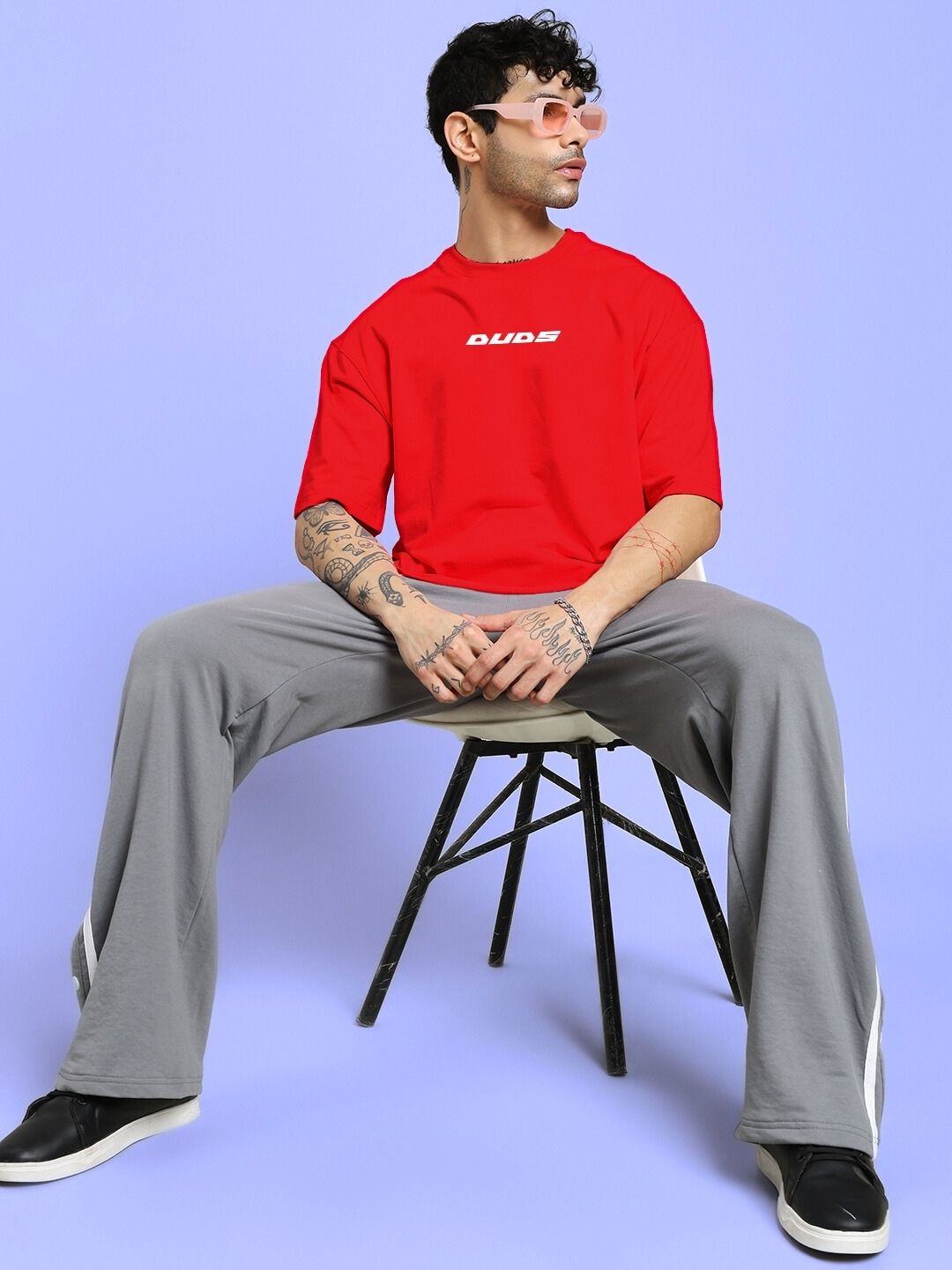 Model wearing CYBERPUNK OVER-SIZED T-SHIRT RED seated against a light blue background, styled with gray pants and sunglasses.