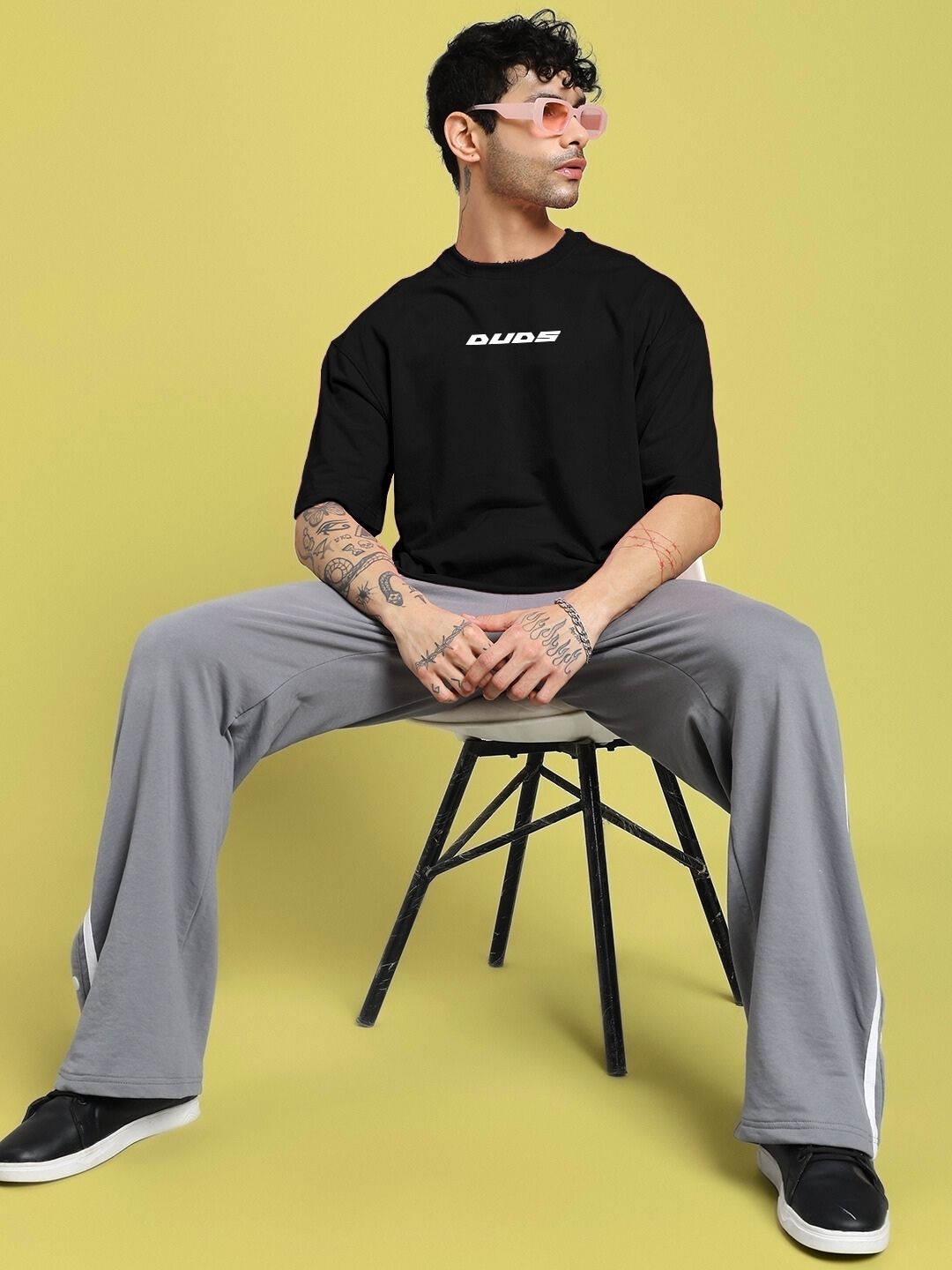 Model wearing CYBERPUNK OVER-SIZED T-SHIRT BLACK with gray pants, sitting on a chair against a yellow background.
