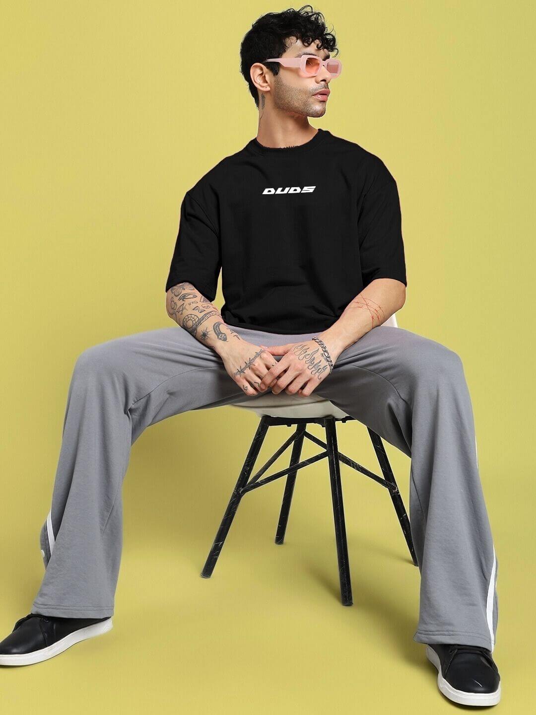 Model wearing CYBERPUNK OVER-SIZED T-SHIRT BLACK with futuristic sunglasses, seated on yellow background
