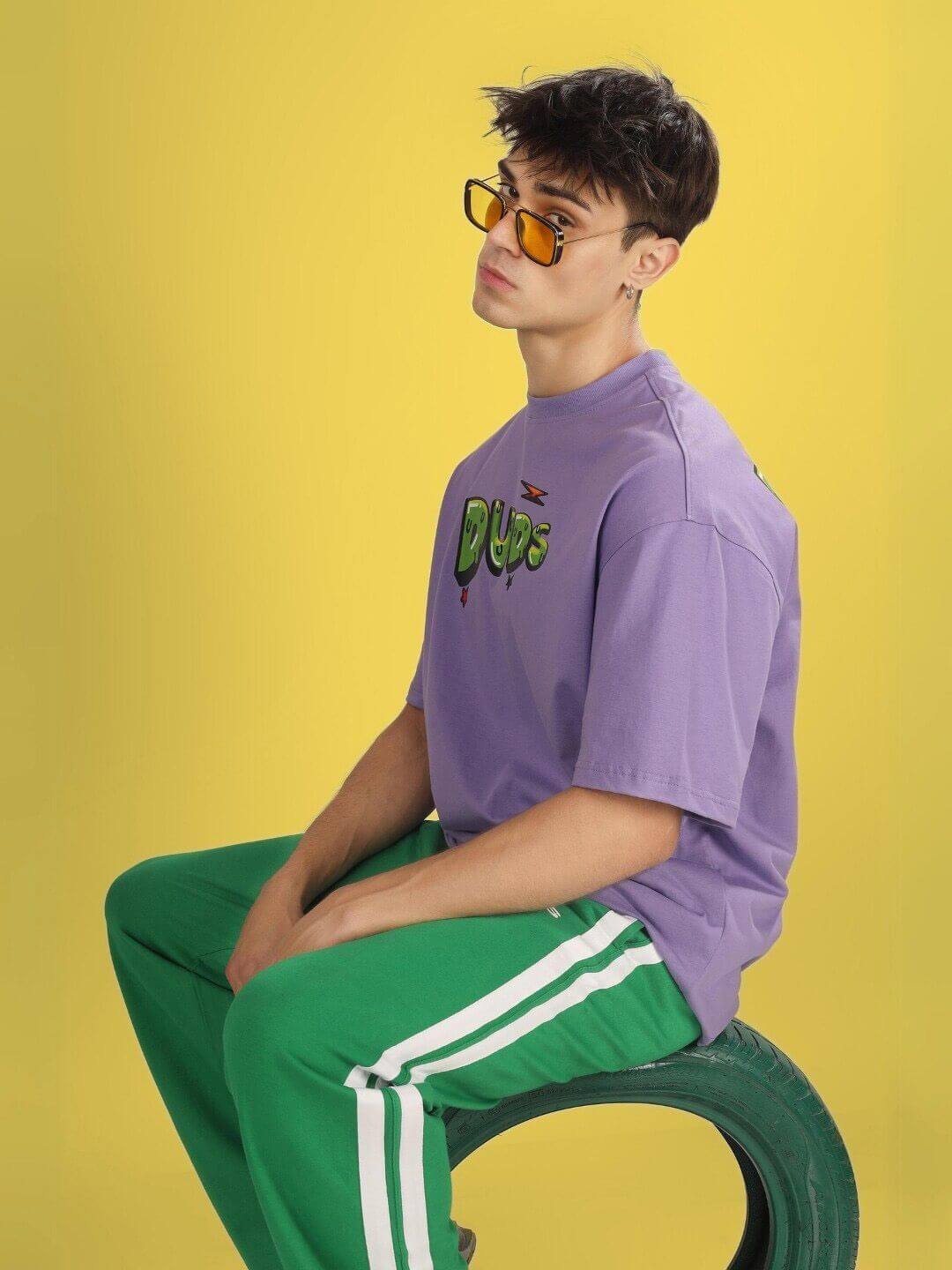 Model wearing ESCAPED TWEETY OVER-SIZED T-SHIRT WISTERIA PURPLE, sitting on tire, yellow background, sunglasses, casual style.