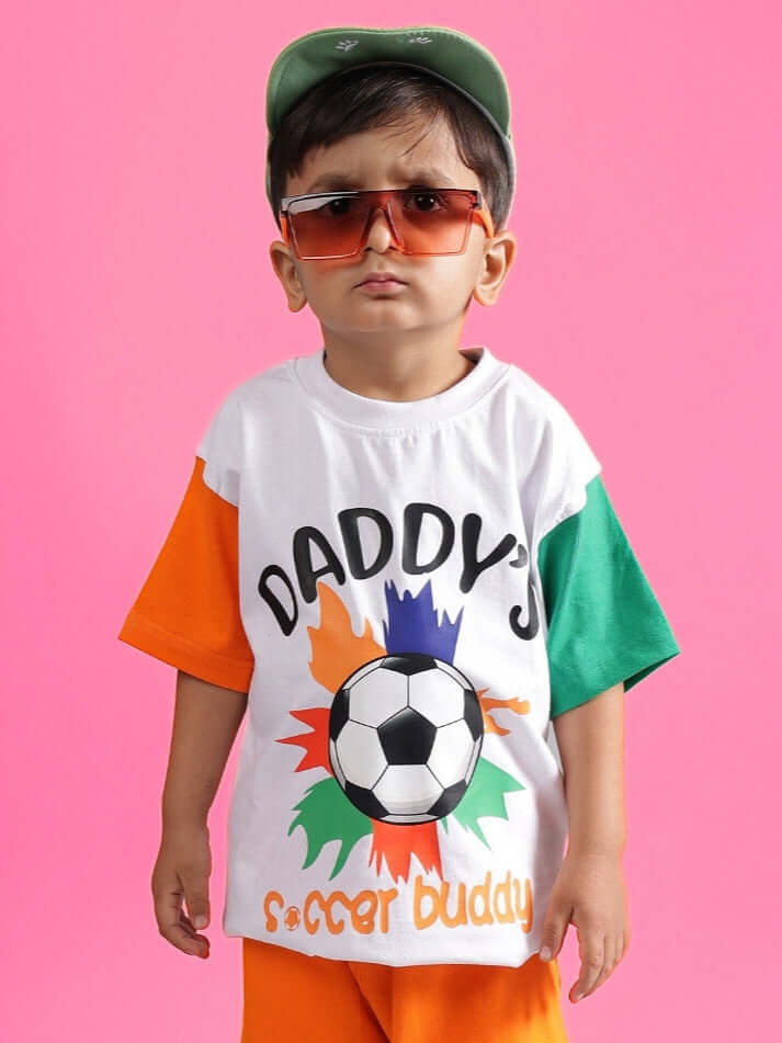 Child wearing a colorful oversized t-shirt with soccer design, sunglasses, and cap, standing against a pink background.