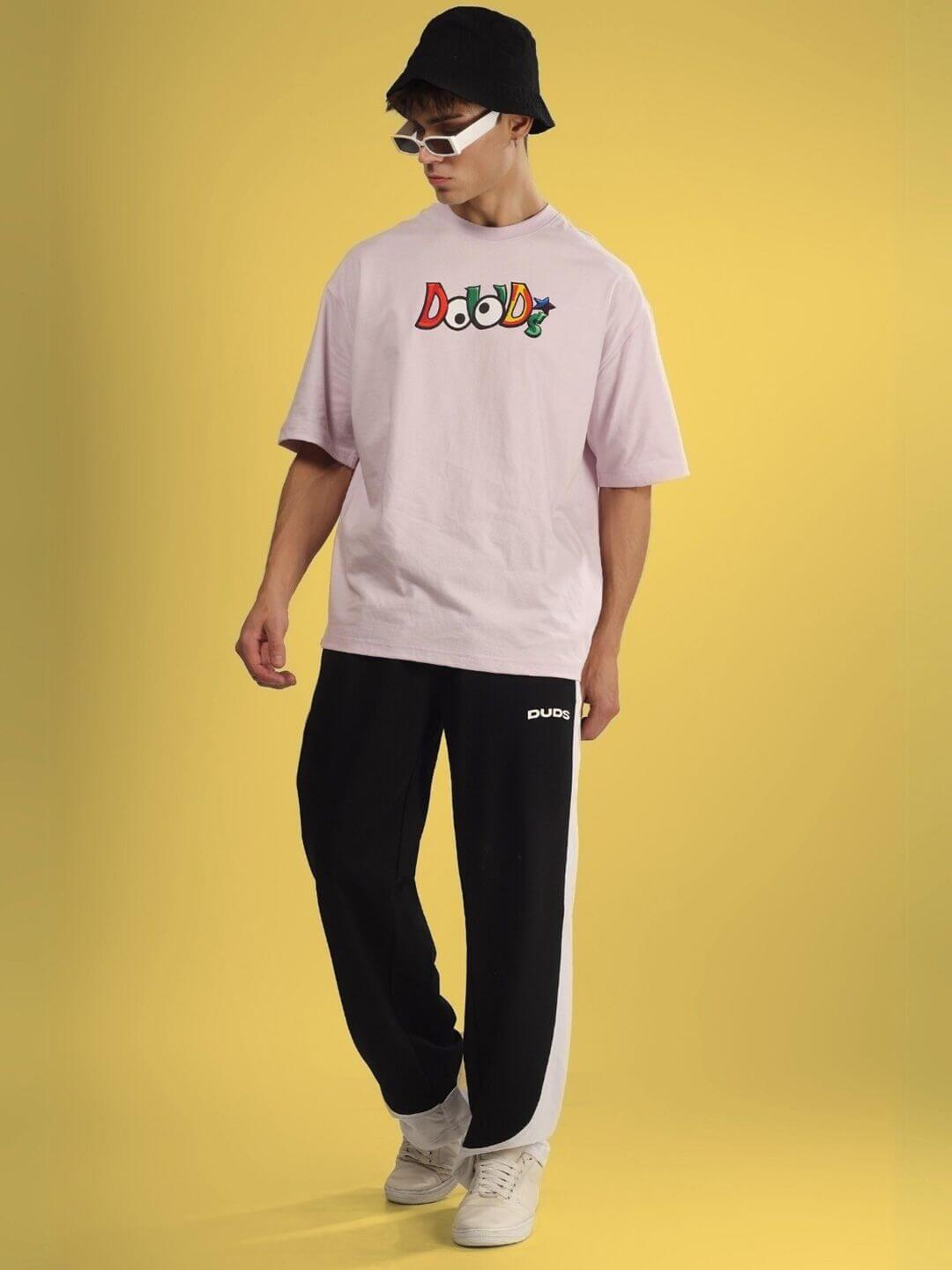 Man wearing DUDS MONSTER OVER-SIZED T-SHIRT PALE PINK with black pants and white sneakers against a yellow background
