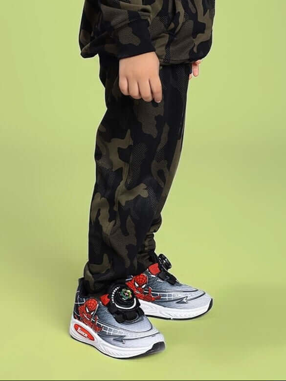 Child wearing DEFENCE JOGGER FOR BOYS AND GIRLS CAMO GREEN, showcasing camouflage pattern and sneakers against a green background.