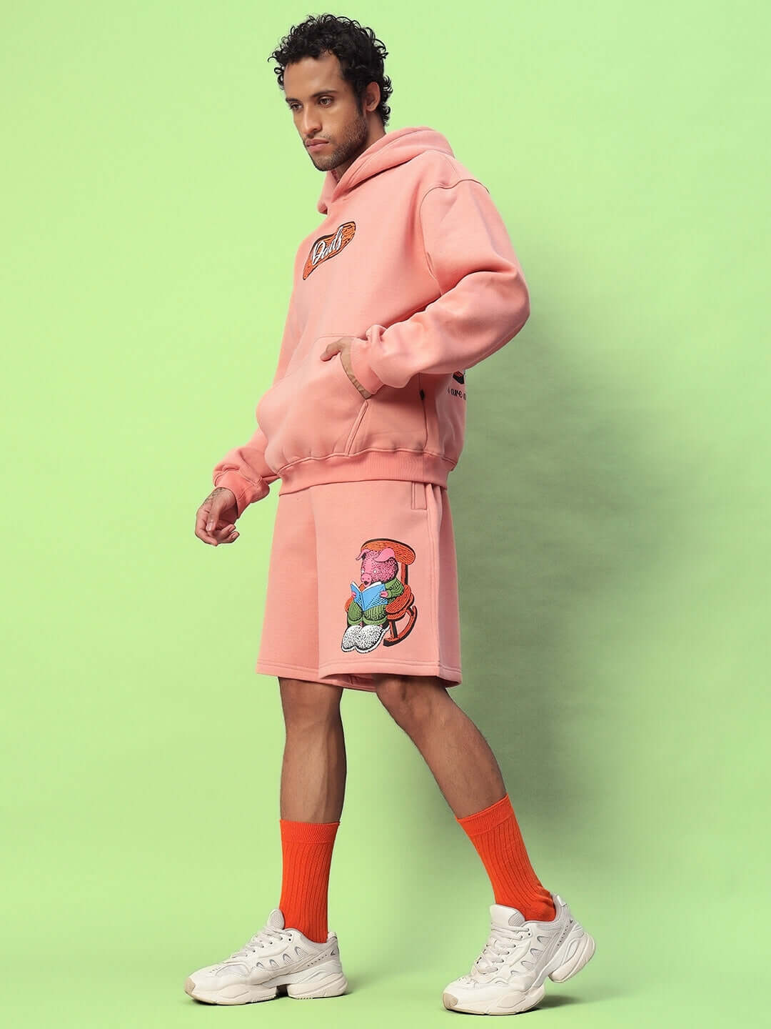 Man wearing Deliberate Fleece Co-Ord Peach loungewear set with hoodie and shorts, standing against green background.