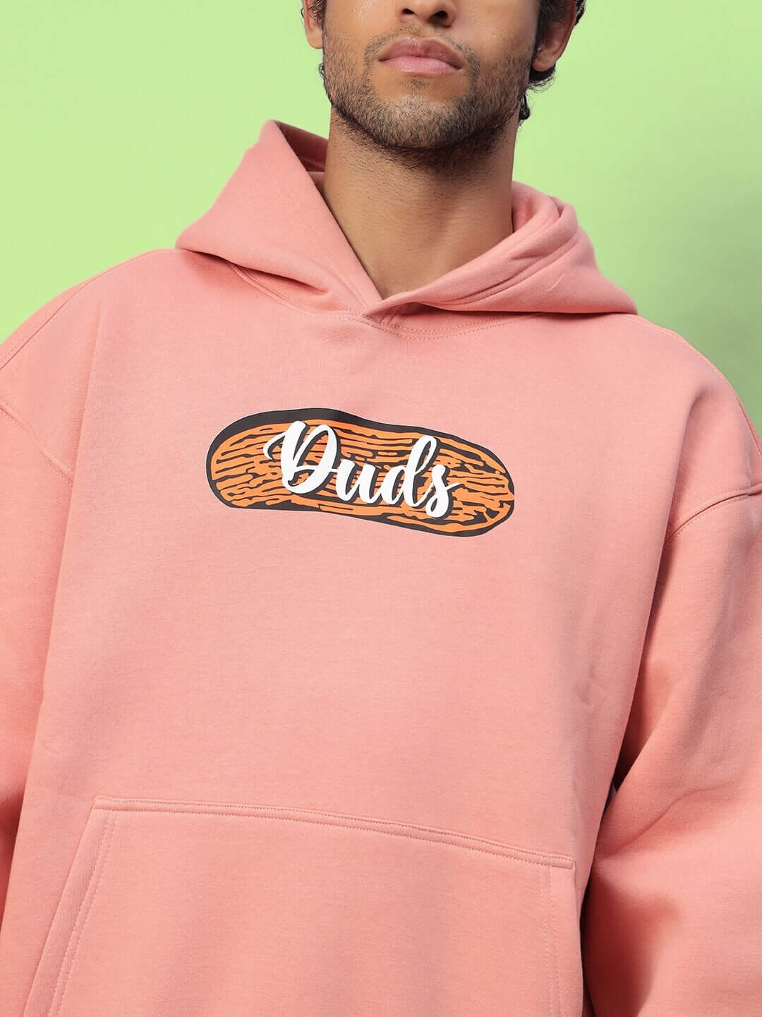 Man wearing Deliberate Fleece Hoodie in peach with graphic text, featuring a relaxed fit and front pocket, set against a green background.