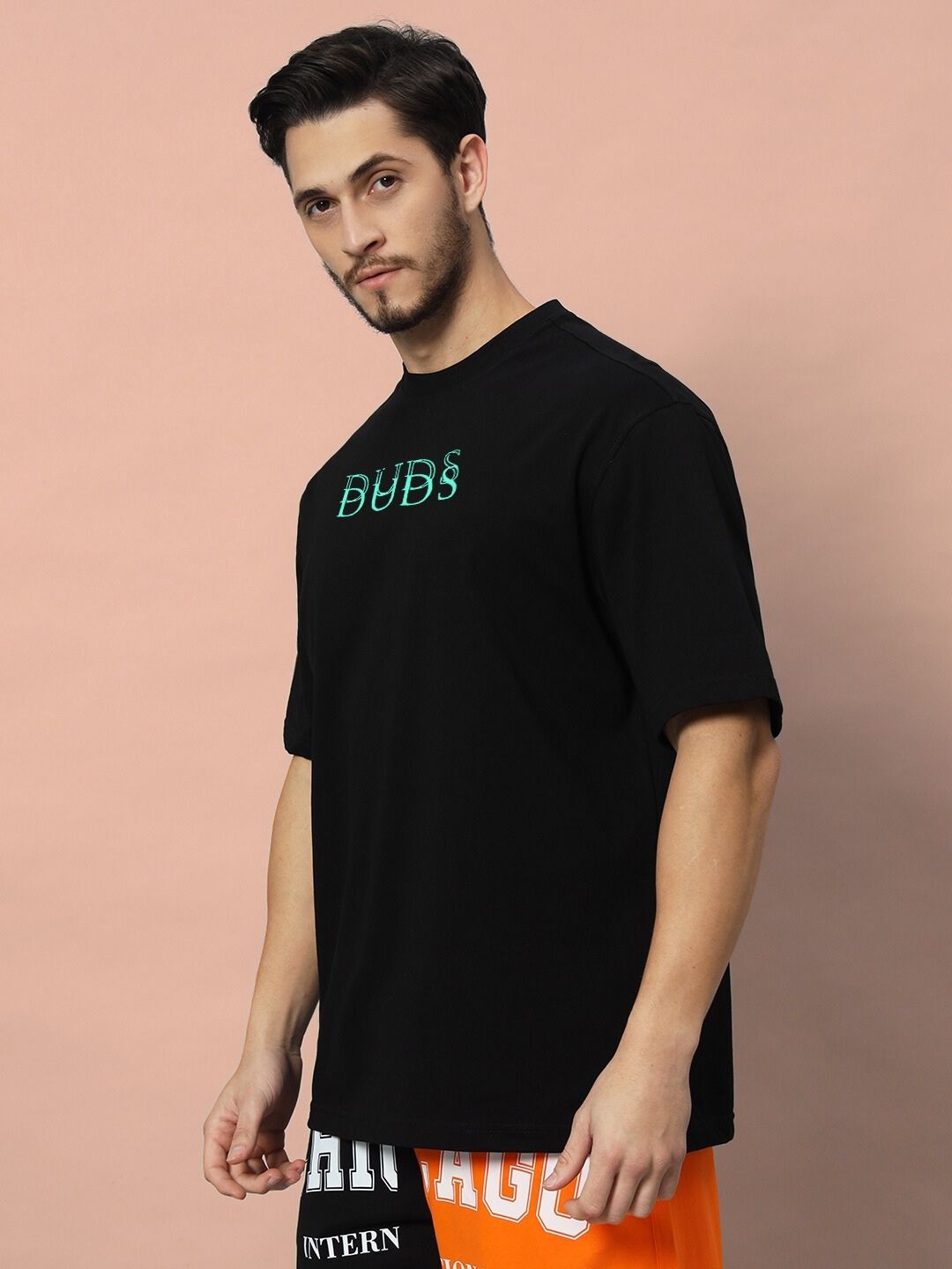 Man wearing DELUSION OVER-SIZED T-SHIRT BLACK with 'BUDS' text on front against a pastel pink background.