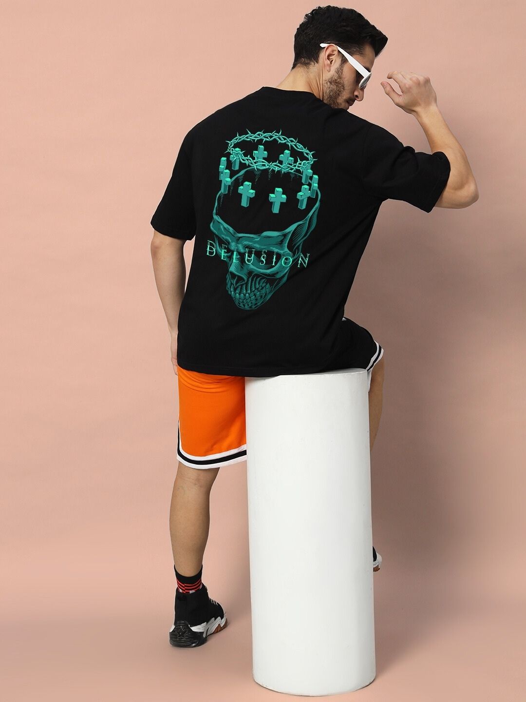 Man wearing DELUSION OVER-SIZED T-SHIRT BLACK with graphic print on back, sitting on white pedestal, facing away from camera.