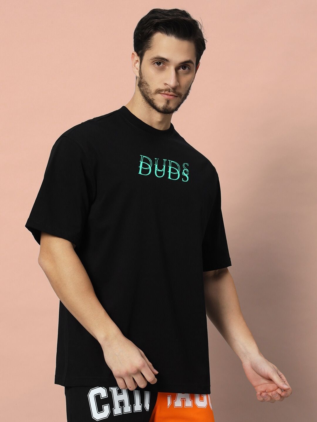 Man wearing DELUSION OVER-SIZED T-SHIRT BLACK standing against peach background, showing front view of t-shirt with green "DUBS" print.