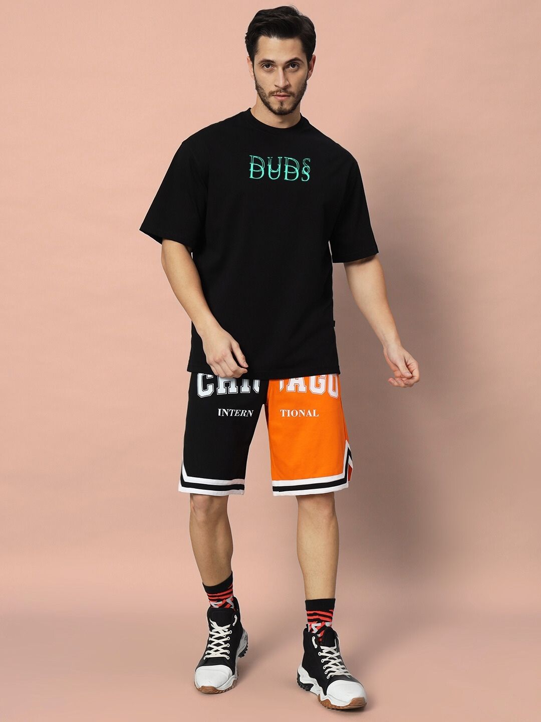 Model wearing DELUSION OVER-SIZED T-SHIRT BLACK standing against a peach background, showcasing casual fashion with black sneakers.