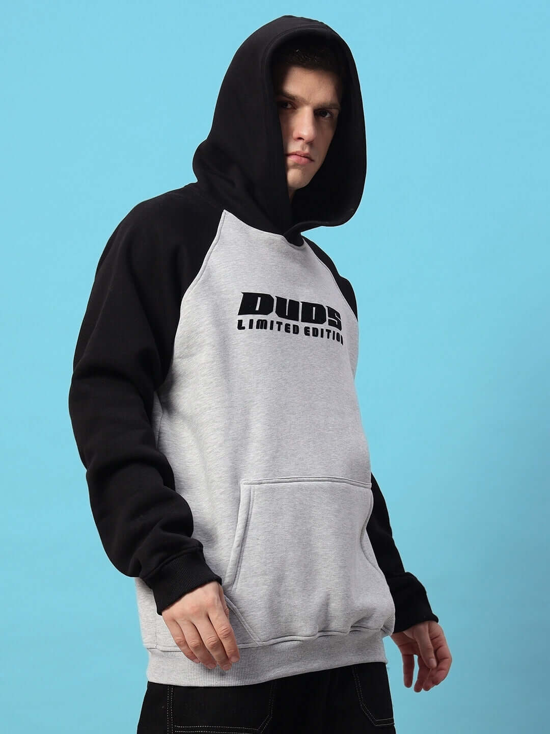 Man wearing DELUXE FLEECE COLORBLOCK OVERSIZE HOODIE BLACK MELANGE GREY, showcasing modern streetwear style against blue background.