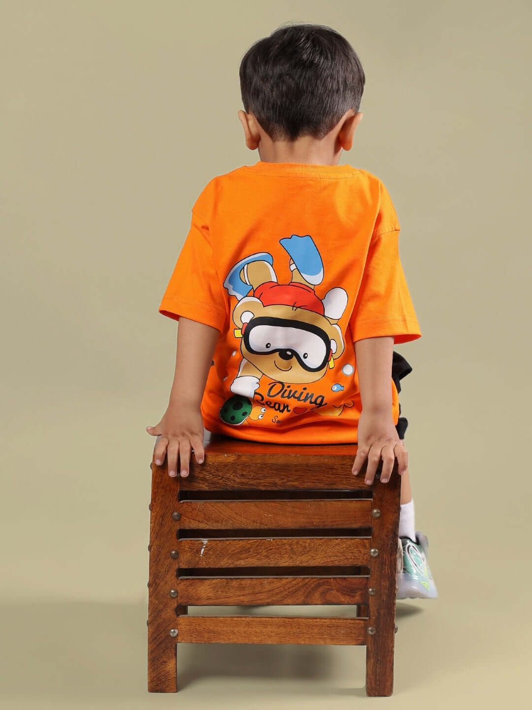 Child wearing DIVING BEAR CO-ORD FOR BOYS & GIRLS ORANGE-BLACK outfit with playful bear design, sitting on a wooden stool.